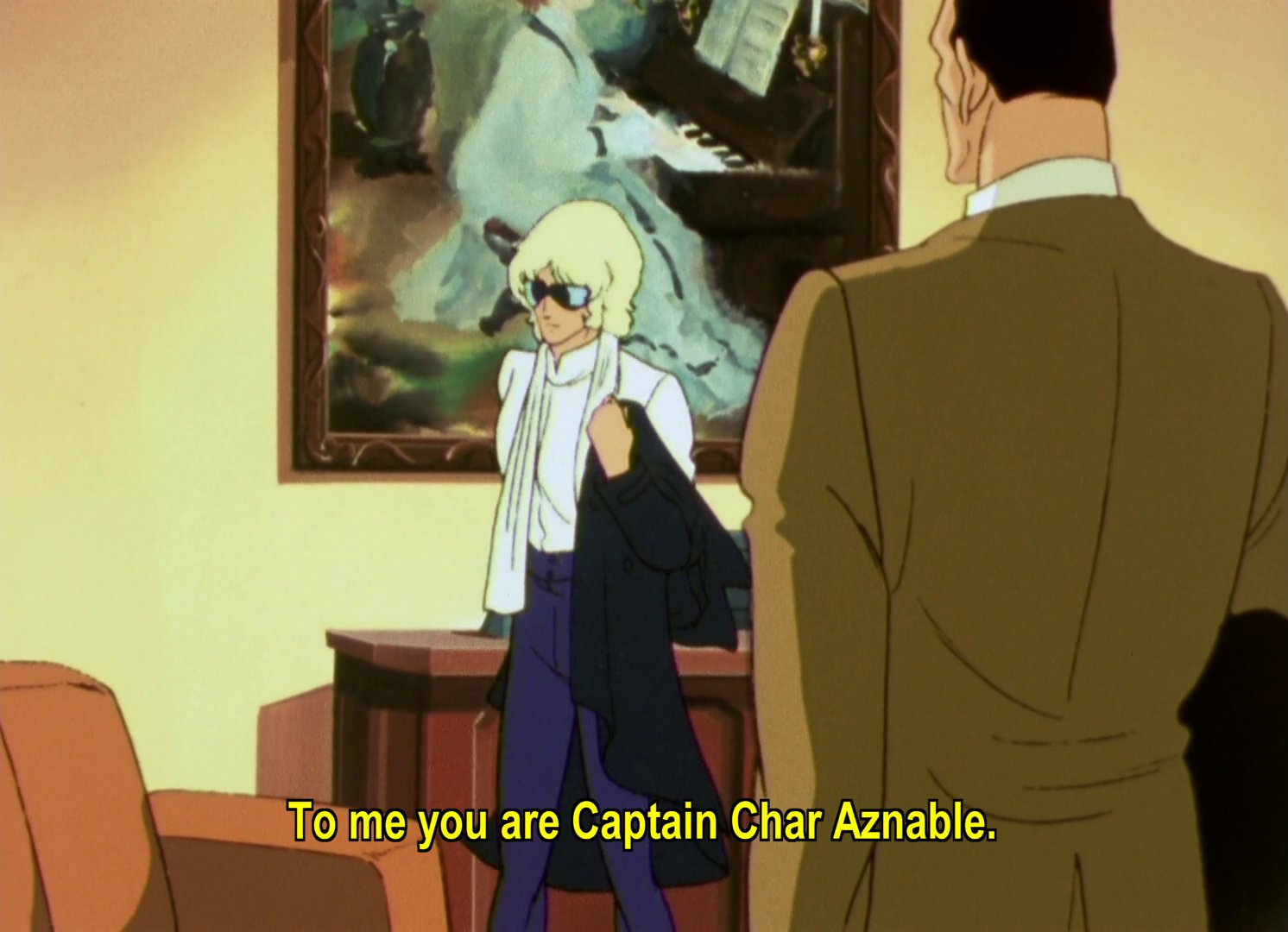 Lt Bajeena in a fancy office, wearing a white shirt, high waisted navy trousers, a long black jacket, and a white scarf.